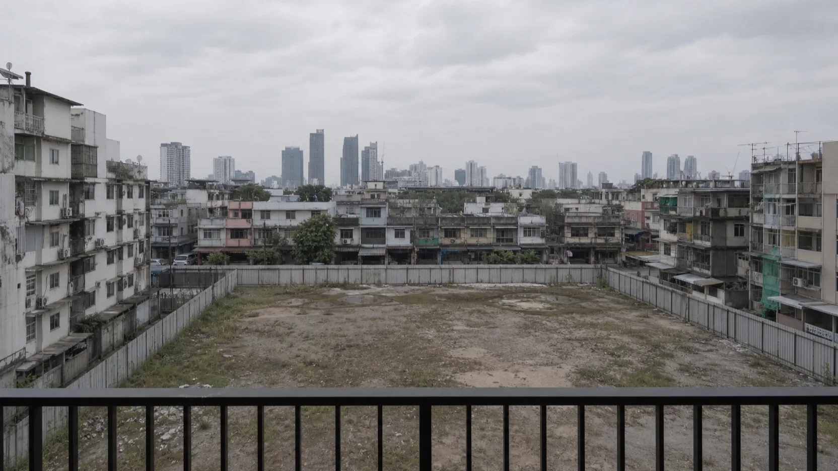 A view study from a mid-floor south-facing unit at NODE Rama 9 — the covered terrace rail in the foreground, the low-rise shophouses and the hoarded vacant plot below, the older buildings and a scatter of towers beyond. Overcast, unflattered.